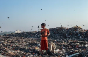 Cette image montre l'inverse de la mode éthique. En effet, un enfant est au milieu d'une très grande décharge de déchets, en majorité textile. Il tient un déchet à la main et des oiseaux naviguent dans le ciel.