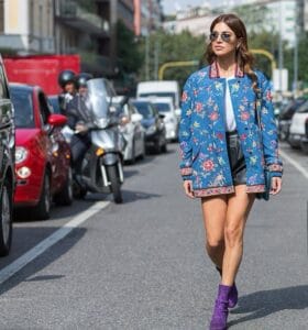 Femme marchant dans une rue animée de Paris, portant une veste bleue à motifs floraux, un short noir et des bottines violettes pailletées, incarnant le style streetwear chic de la Fashion Week.