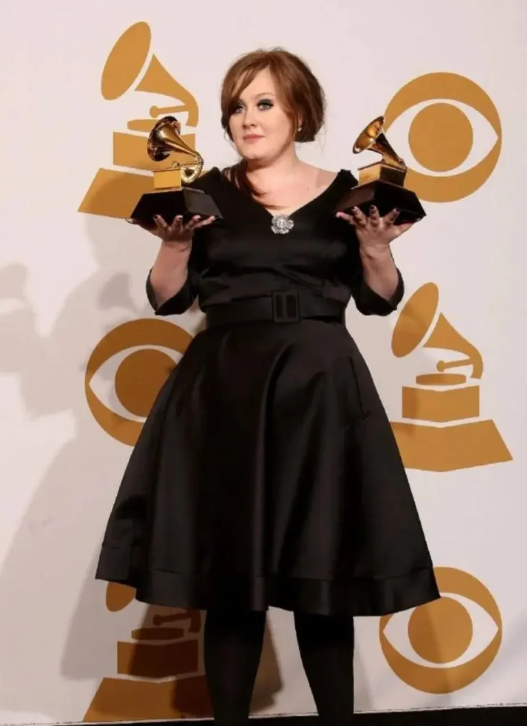 Femme en robe de soirée noire tenant deux trophées de couleur dorée devant un fond doré avec des logos de grammy.