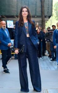L'image montre une femme aux longs cheveux bruns posant avec élégance devant un groupe de personnes, vêtue d'un costume bleu marine composé d'une veste et d'un pantalon large.