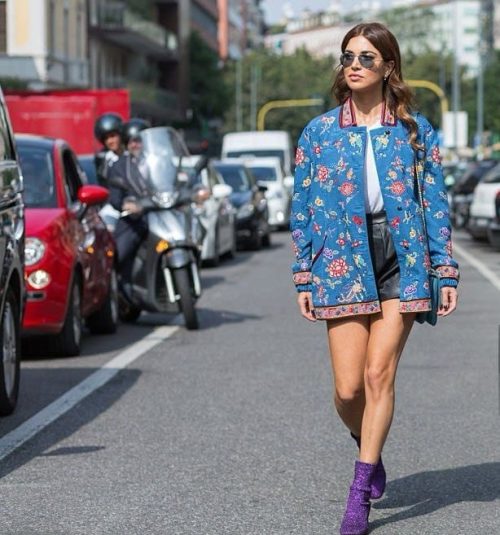 Street Style de la Fashion Week Paris 2025 : Entre Audace et Élégance Femme marchant dans une rue animée de Paris, portant une veste bleue à motifs floraux, un short noir et des bottines violettes pailletées, incarnant le style streetwear chic de la Fashion Week.